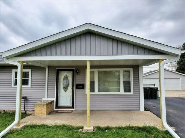 Gray-sided house with a covered porch. White trim, door, and windows. Concrete porch and guttering. Cloudy sky.