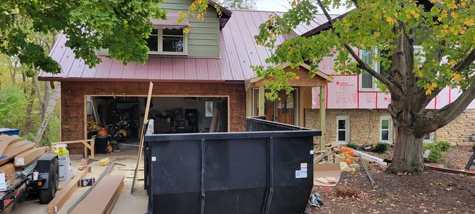 House under construction with a large dumpster in front. Garage door open. Fall foliage.