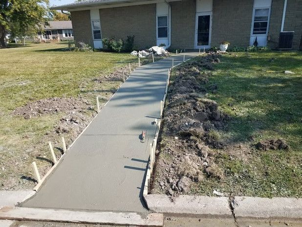 New concrete sidewalk being poured in front of a brick house. Stakes support the formwork.