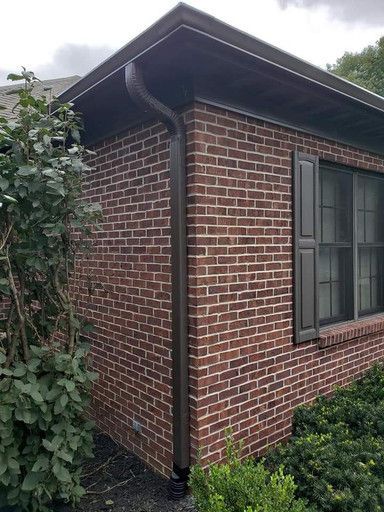 Corner of a brick building with dark gutters and a window with brown shutters, surrounded by bushes.