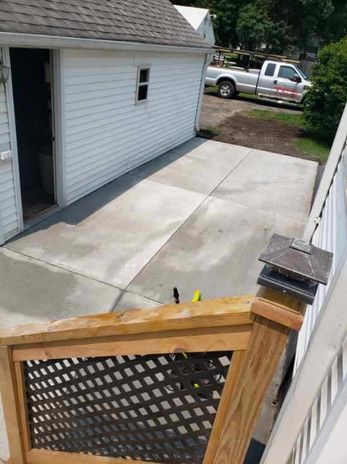 Concrete patio next to a white garage, viewed from a wooden deck with railing. A pickup truck is in the background.