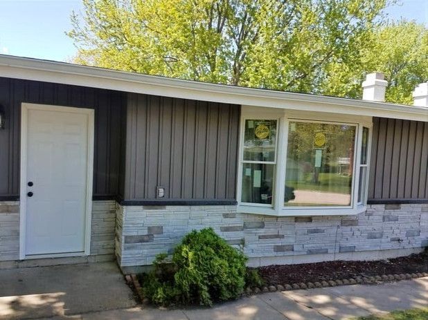 Exterior of a house with gray siding, white door, and bay window. Decorative stone on lower portion.
