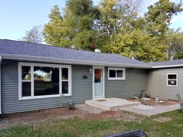 Gray-sided ranch house with large windows, light blue door, and small concrete patio. Green trees in the background.