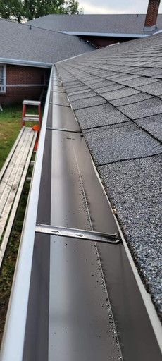 A close-up view of a roof gutter. The silver gutter is mounted on a gray shingled roof.