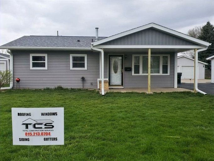 Gray house with light siding, a covered porch, and a sign for roofing, siding, and gutters in the yard.