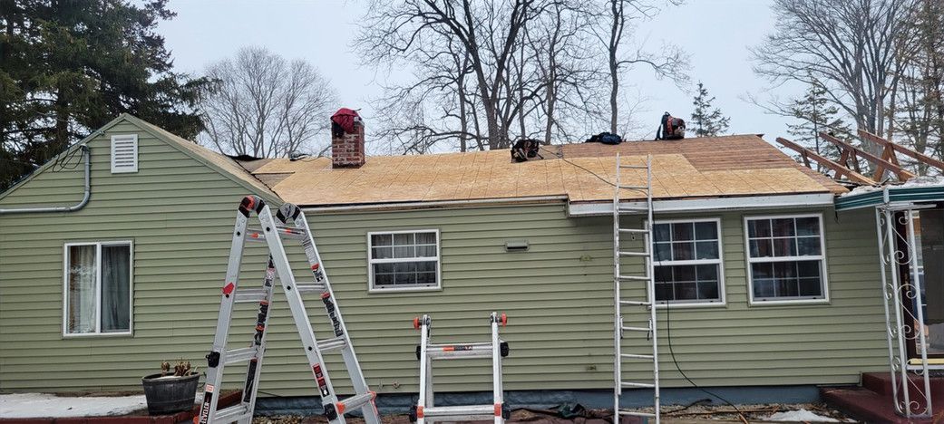 House roof being replaced with workers on the roof, ladders leaning against the house, and trees in the background.