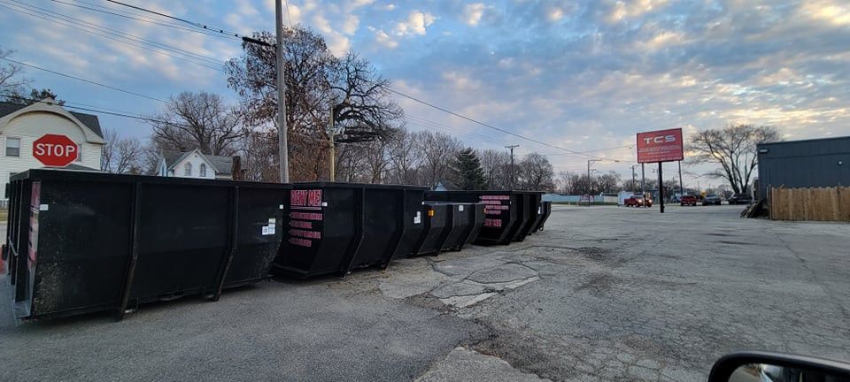 Dumpsters lined up in a lot, in front of a red sign and stop sign under a cloudy sky.