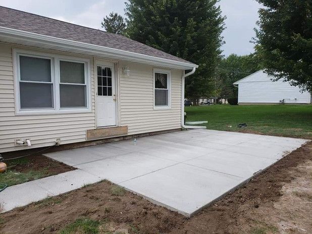 Concrete patio next to a beige house with a white door. Green grass and trees are in the background.