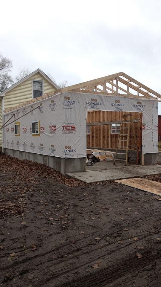 Garage under construction, wood framing and tarp-covered walls with a residential building in the background.