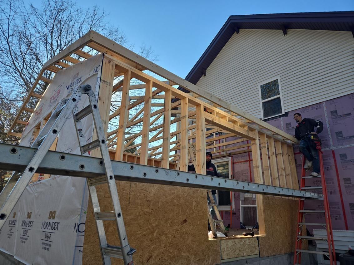 Construction of an addition to a house; workers on ladders framing walls and roof, using lumber.