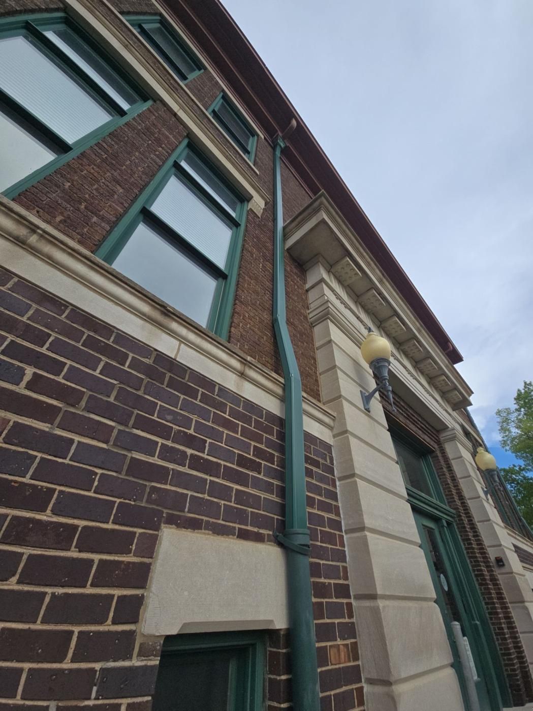 Brick building with green trim, windows, and downspout against a cloudy sky.