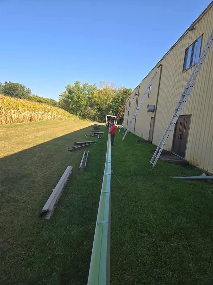 Man working on a long, green gutter along a building's side on a grassy area, with a ladder.