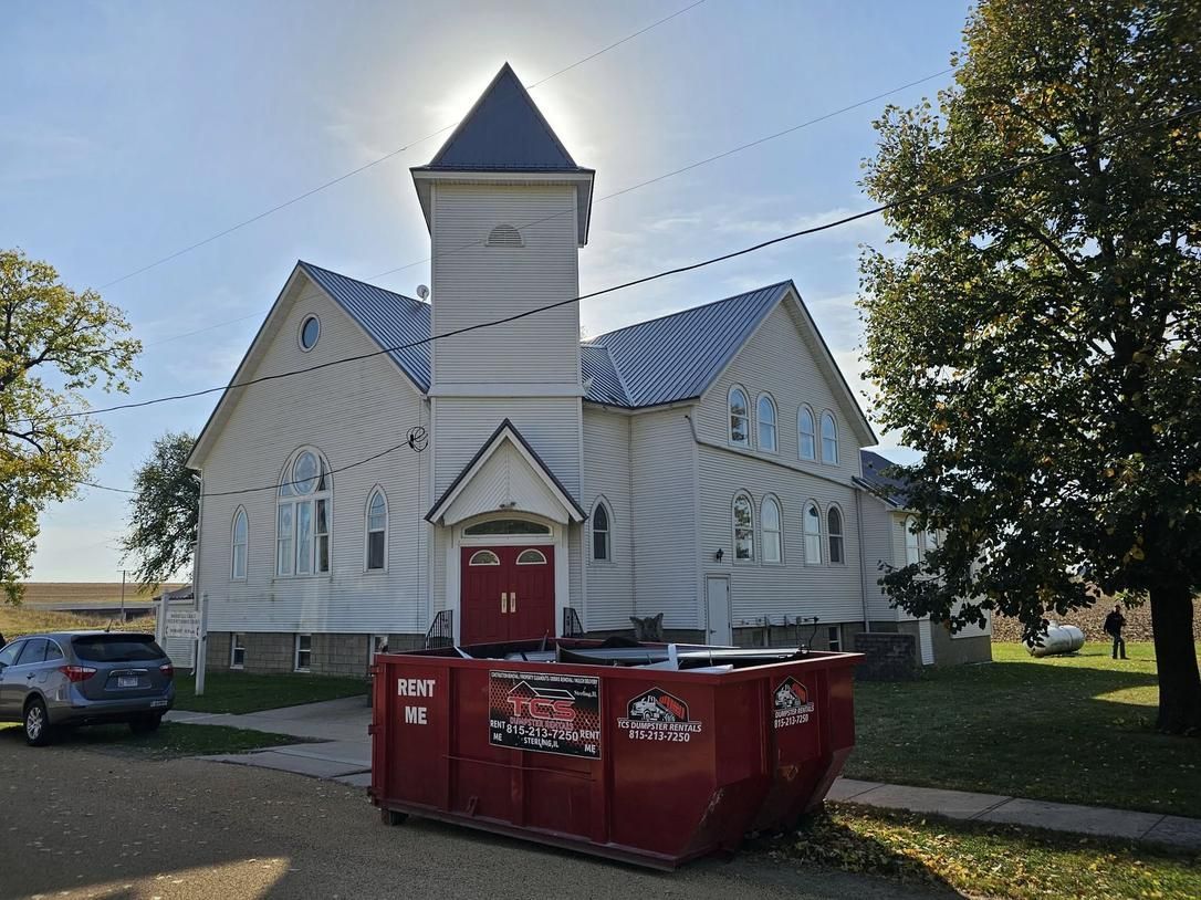 Red dumpster in front of a white church with a tower on a sunny day.