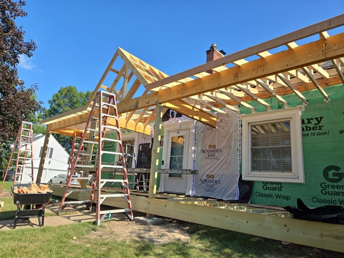 House under construction; wooden roof framing and porch. Exterior covered in green waterproof material. Ladders and wheelbarrow visible.