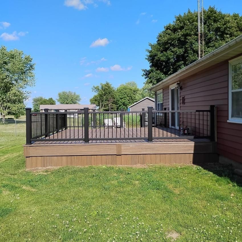 Brown composite deck with black railings next to a brown house with a green lawn and blue sky.