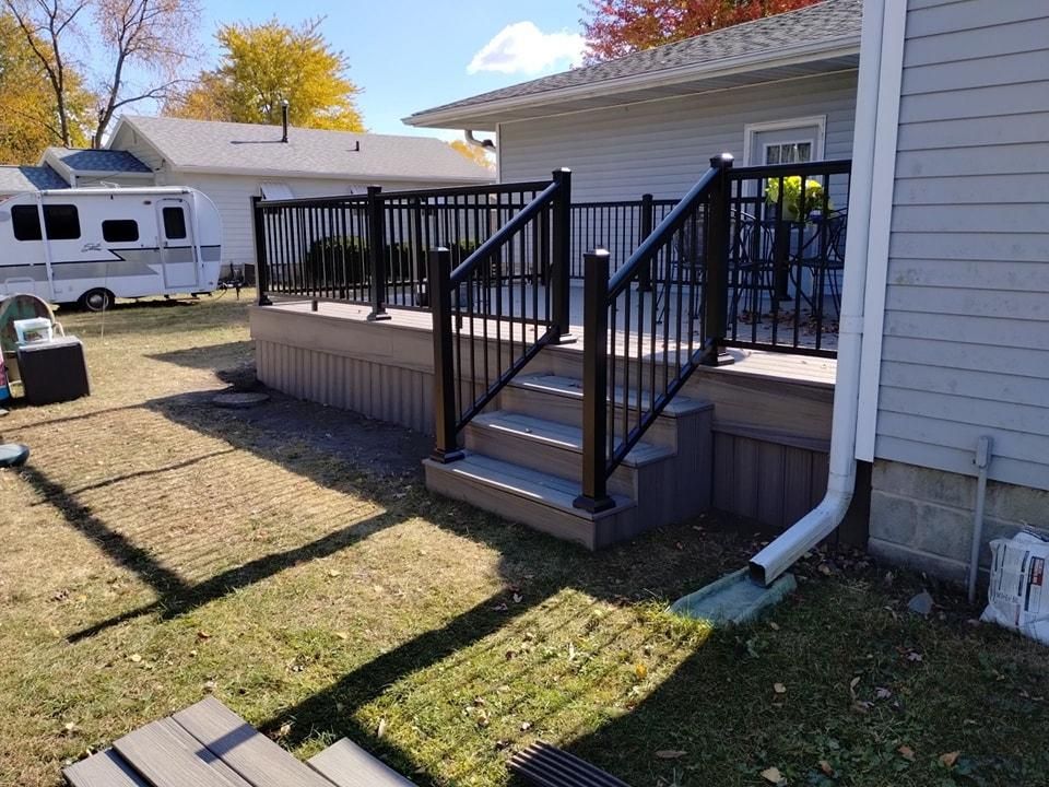 A black-railed deck with stairs, attached to a gray house, set on a green lawn.
