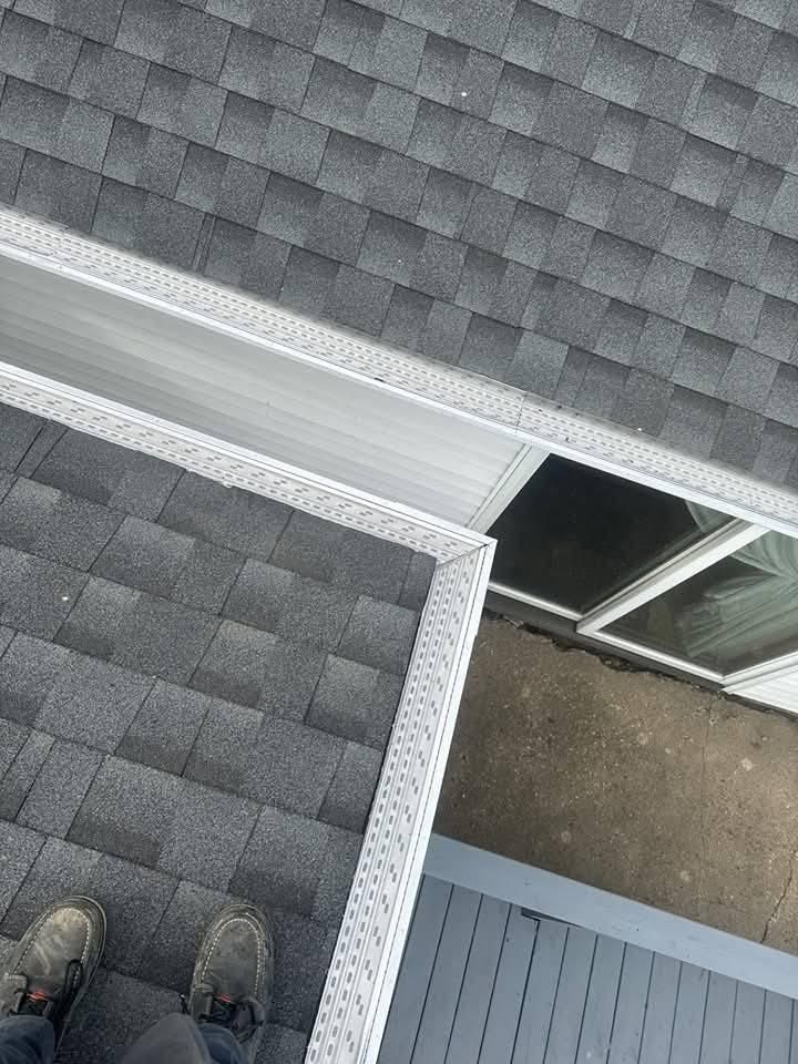 Overhead view of a roof with shingles, gutter, and a window. Someone's feet are visible.
