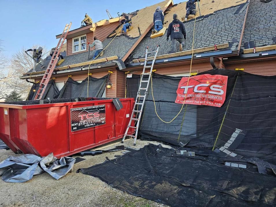 Roofing crew on a house, tearing off old shingles. Red dumpster, safety harnesses, ladders, and black tarps are present.