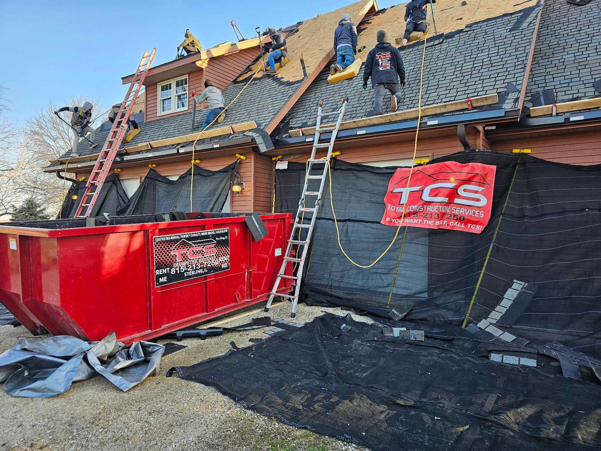Roofing crew replacing shingles on a house. Red dumpster and ladder are visible.