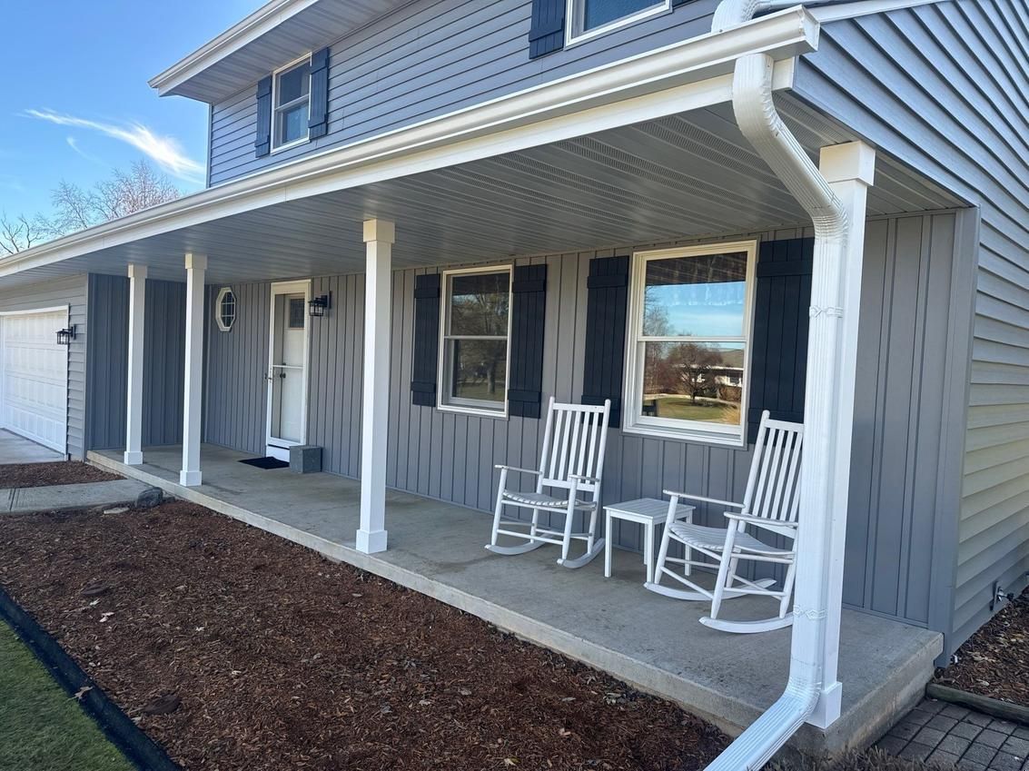 Gray house with a porch. White rocking chairs sit on the porch, a white door, and blue siding.