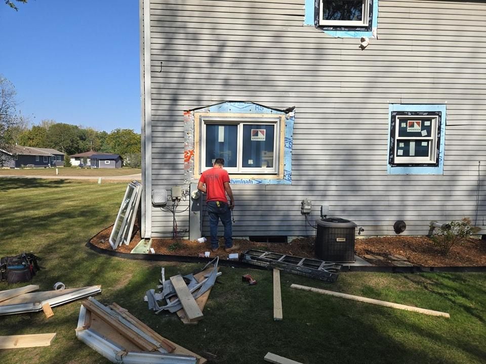 Man installing a window on a house with gray siding; tools and materials are on the grass.