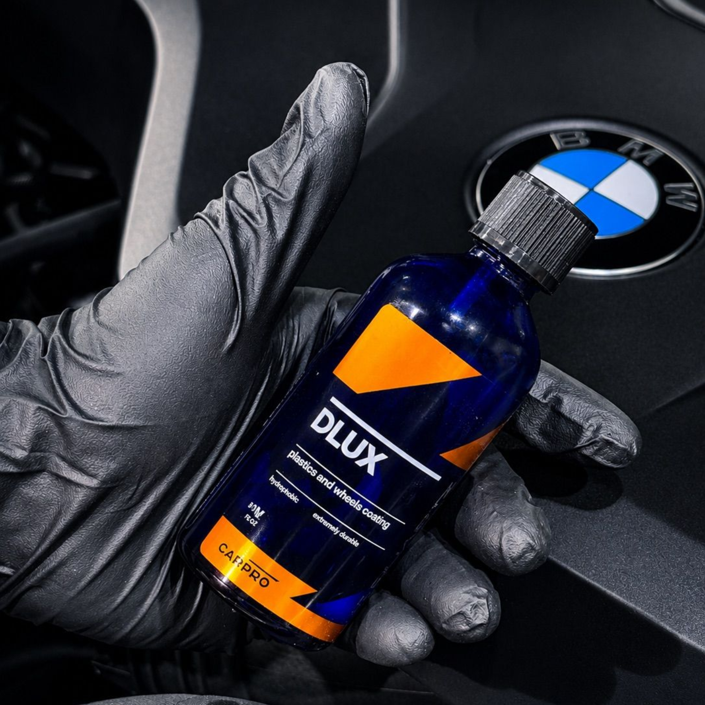 Gloved hand holding a blue Dlux bottle beside a BMW logo on a car dashboard