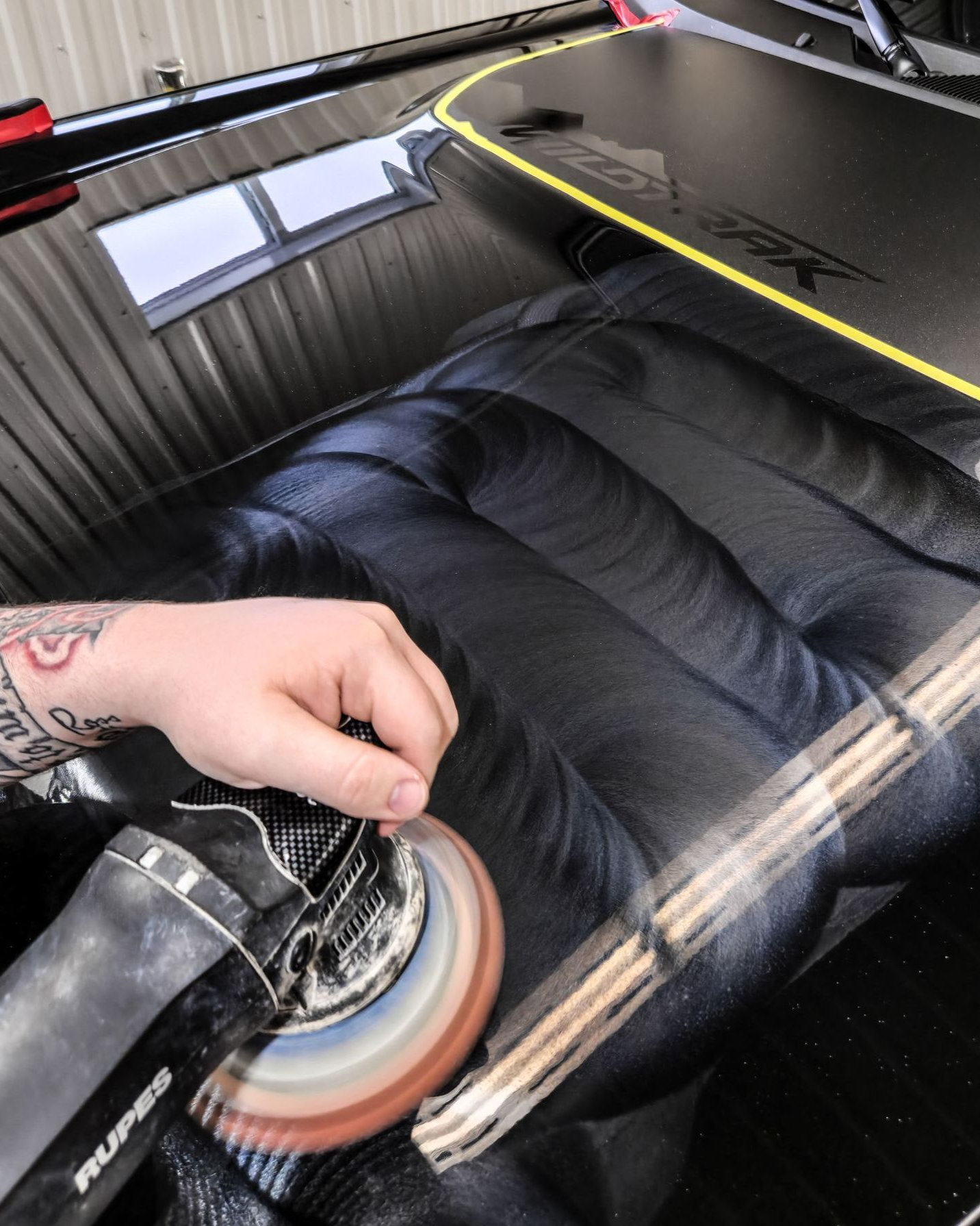 Hand polishing a black car’s body panel with a buffer, reflecting overhead lights.