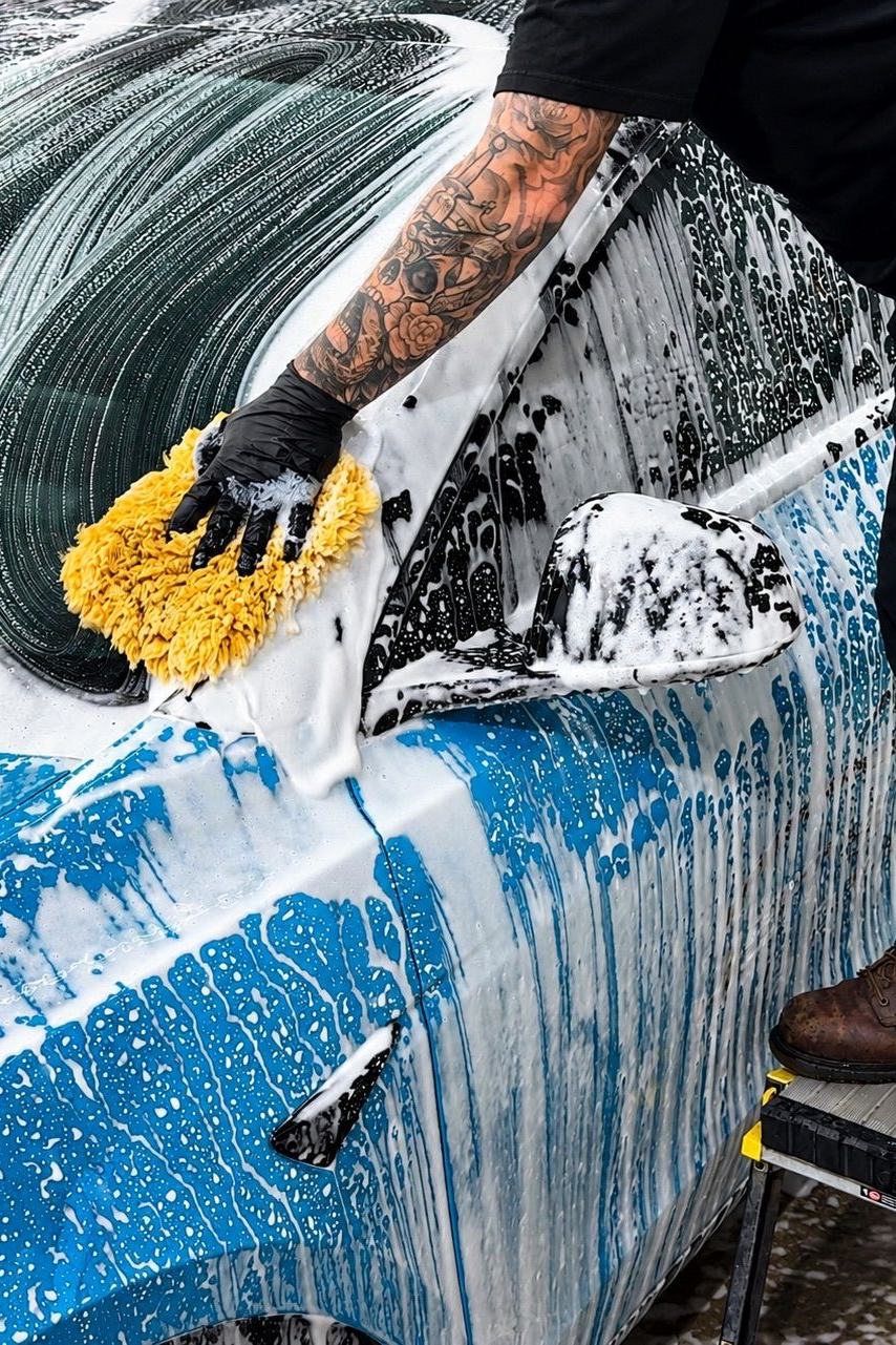 Person washing a blue car with a yellow sponge, covered in soapy suds