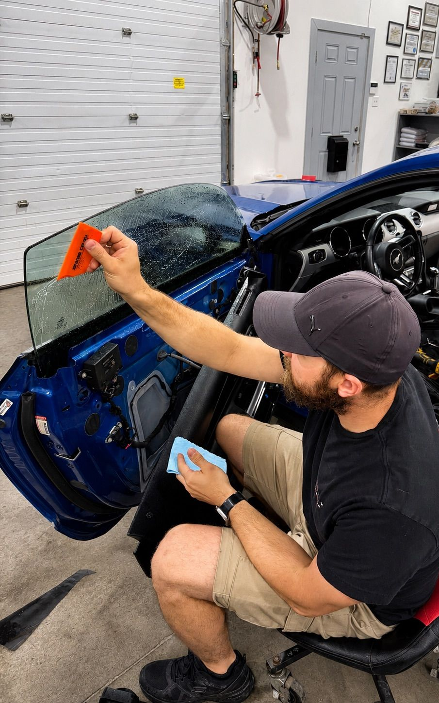 Person applies orange tape to a blue car door in a garage workshop.