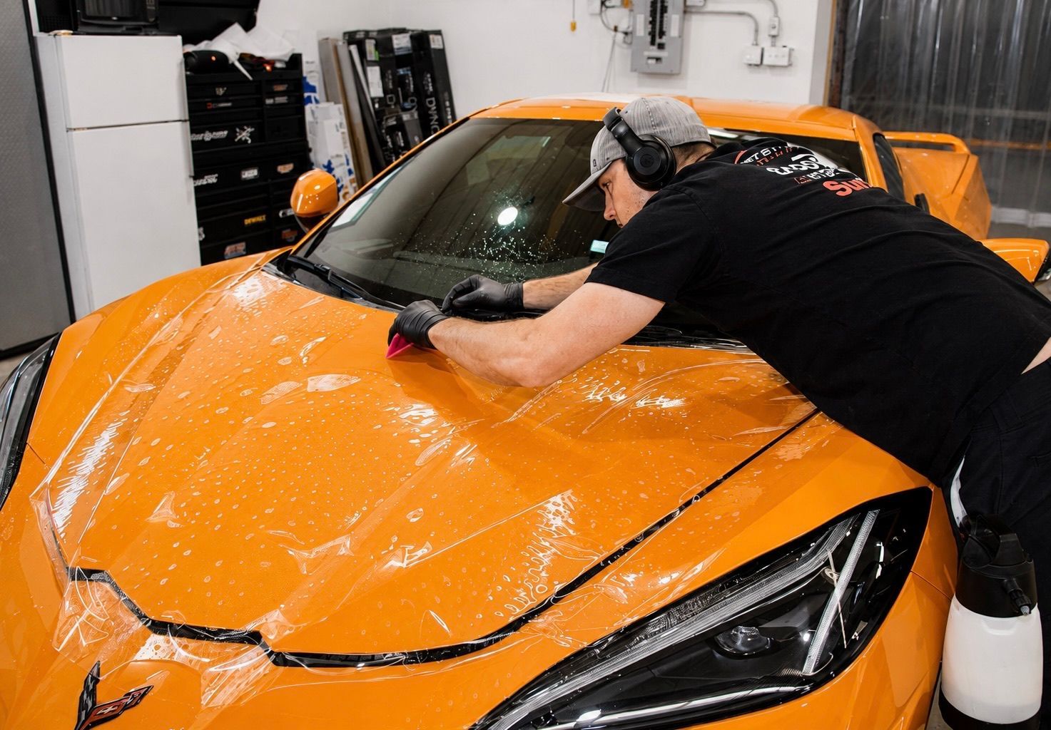 Person polishing an orange sports car in a garage with protective film on the hood