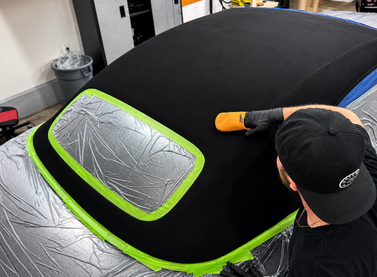 Person polishing a black car roof with green tape and a silver-covered window in a workshop