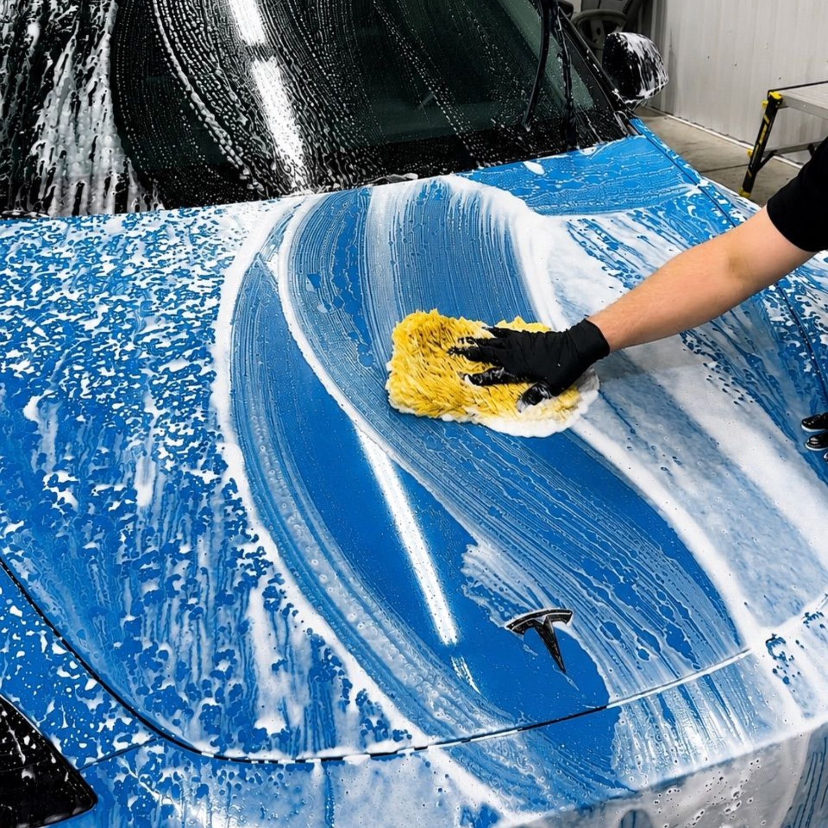 Person washing a blue car hood with a sponge and soapy foam