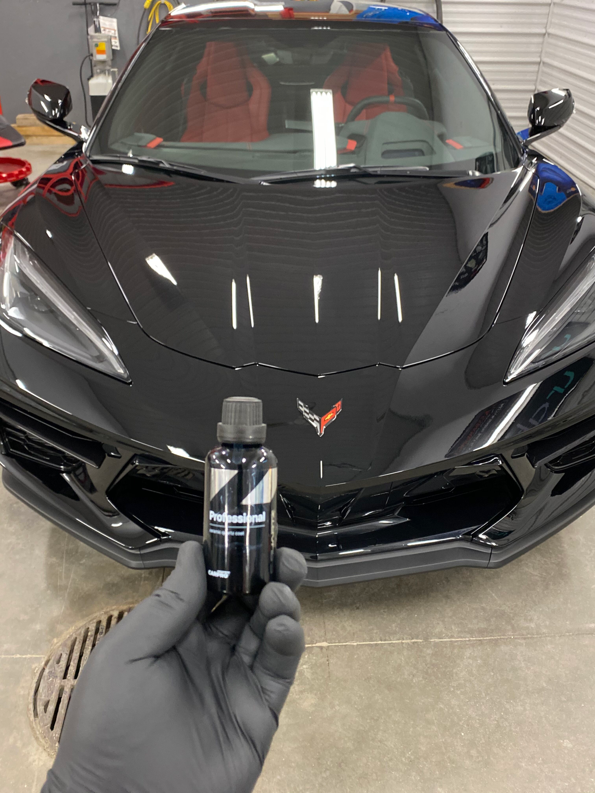 Gloved hand holding a car detailing spray in front of a black Corvette in a garage