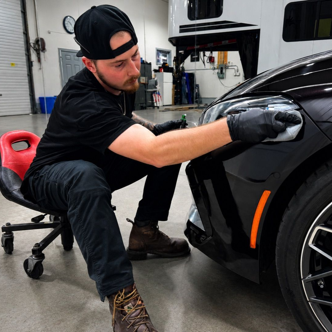 Mechanic polishing a black car’s front fender in a garage, wearing gloves and a cap.