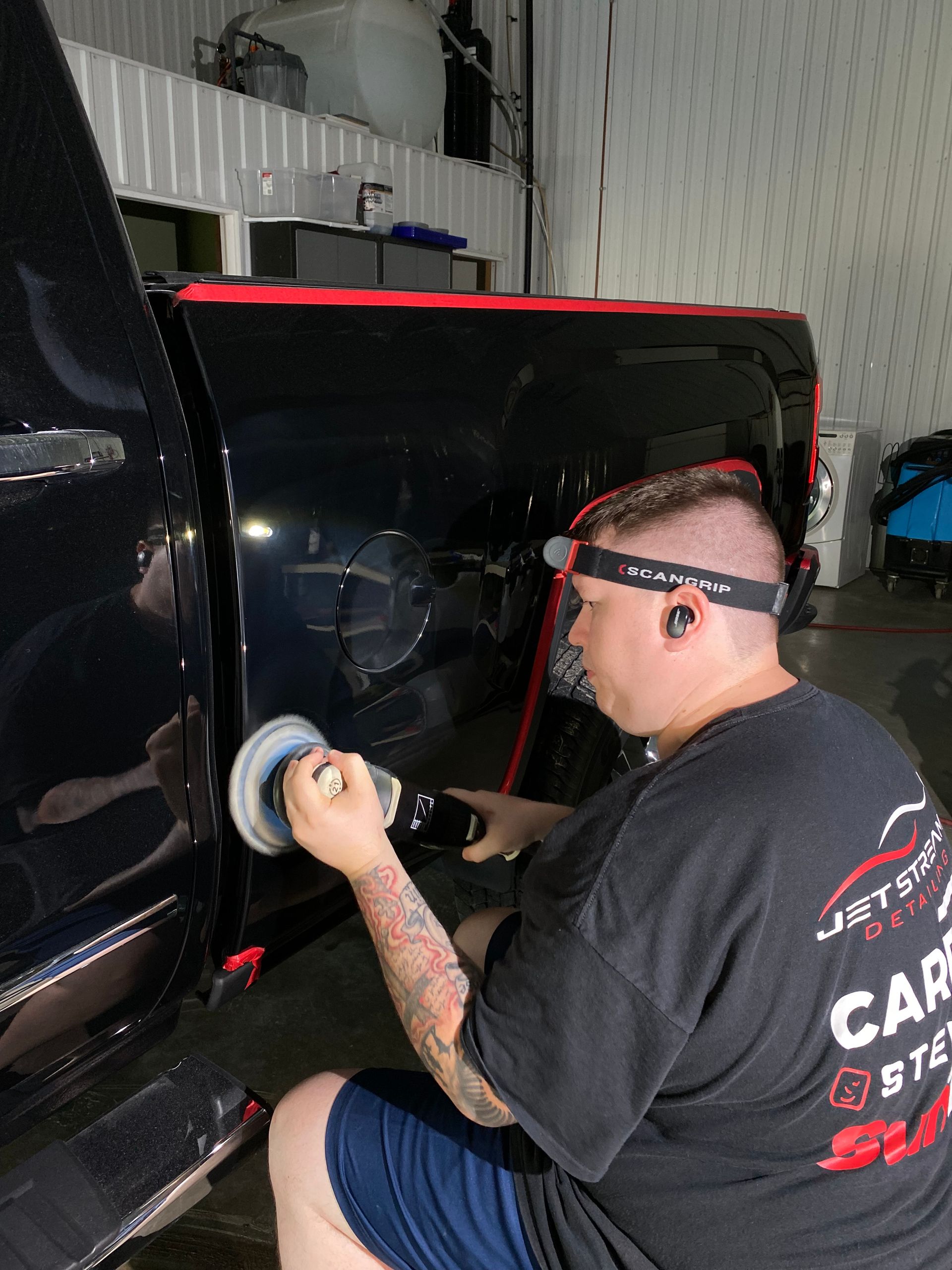 Person polishing a black vehicle panel in a garage, using a handheld buffer near a red roofline.