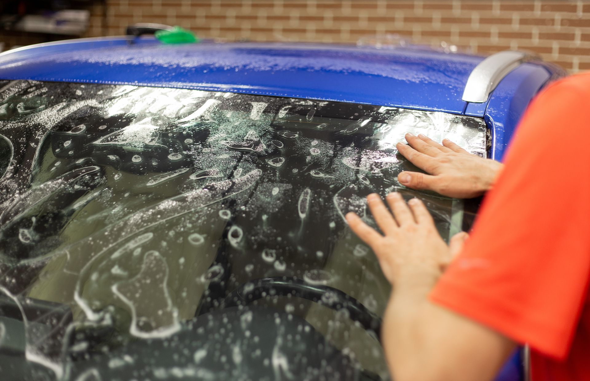 Hands cleaning a blue car windshield with soap suds on the glass