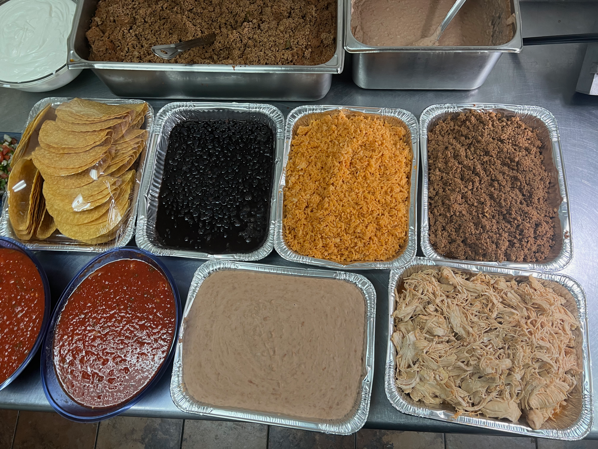 Display of various Mexican food ingredients in metal trays: tortillas, beans, rice, meat, and sauces.