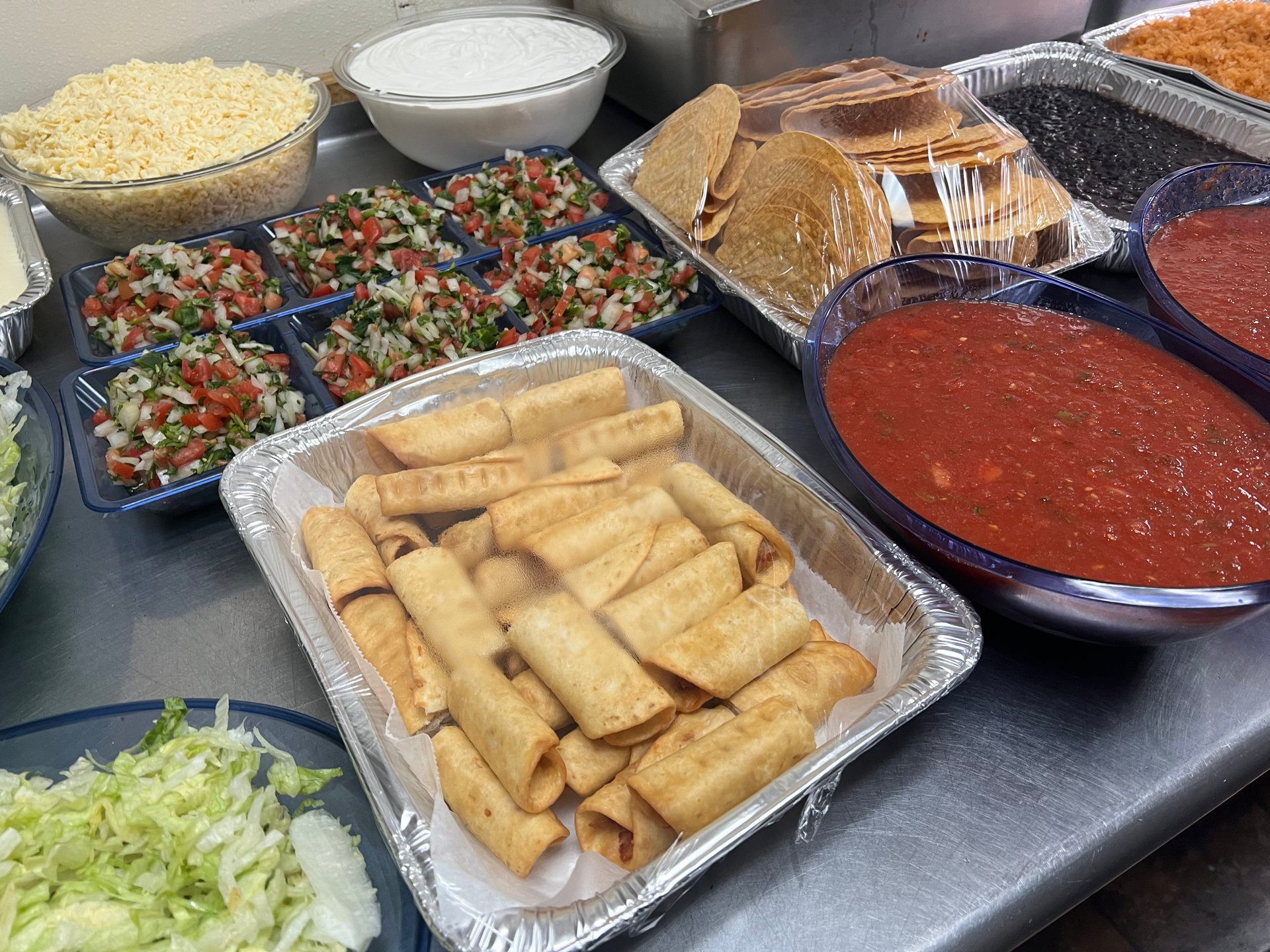 A spread of taco fixings: shredded cheese, salsa, tortillas, taquitos, and more.