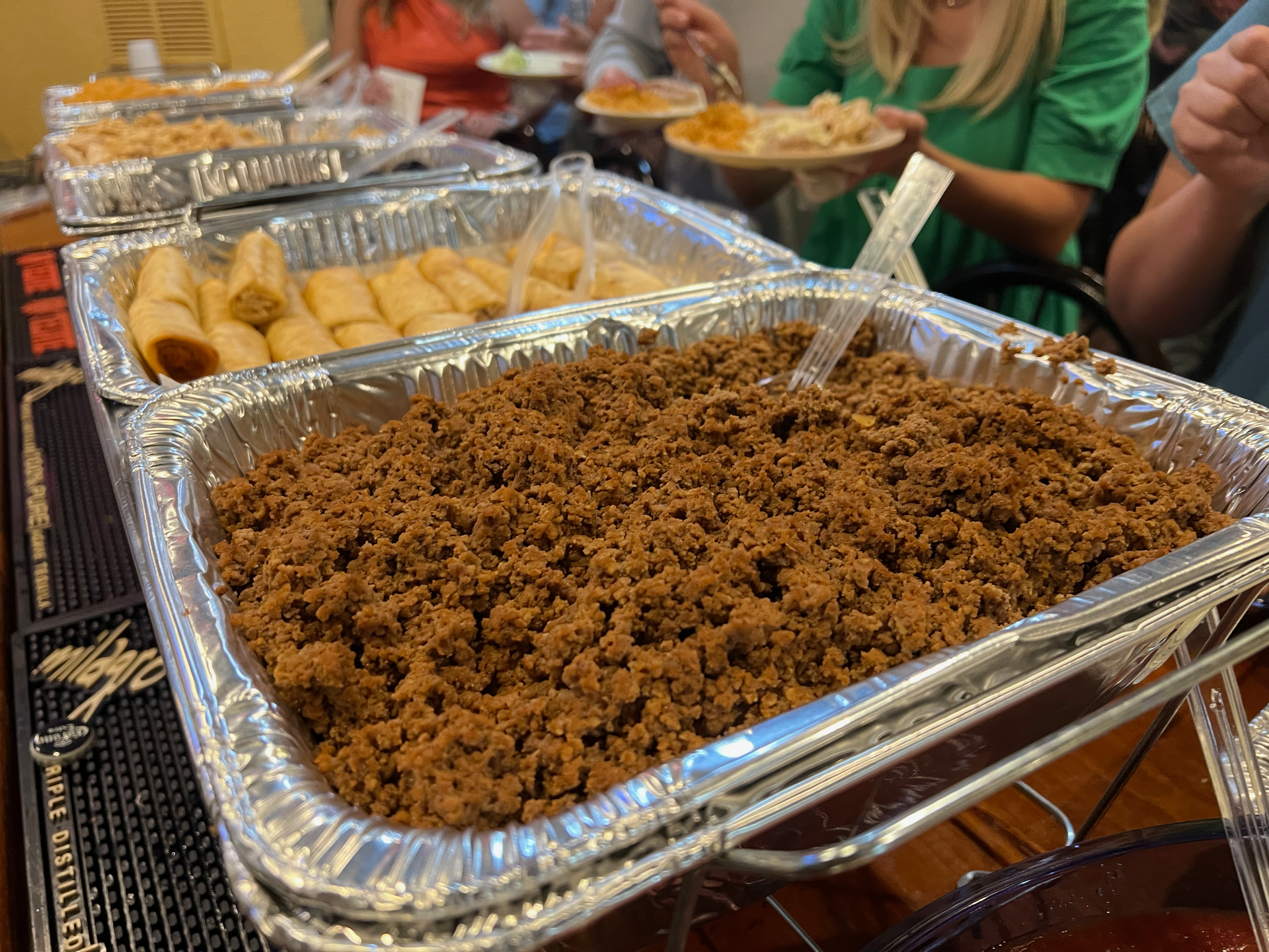 Buffet with trays of ground meat, egg rolls, and other food; people eating.