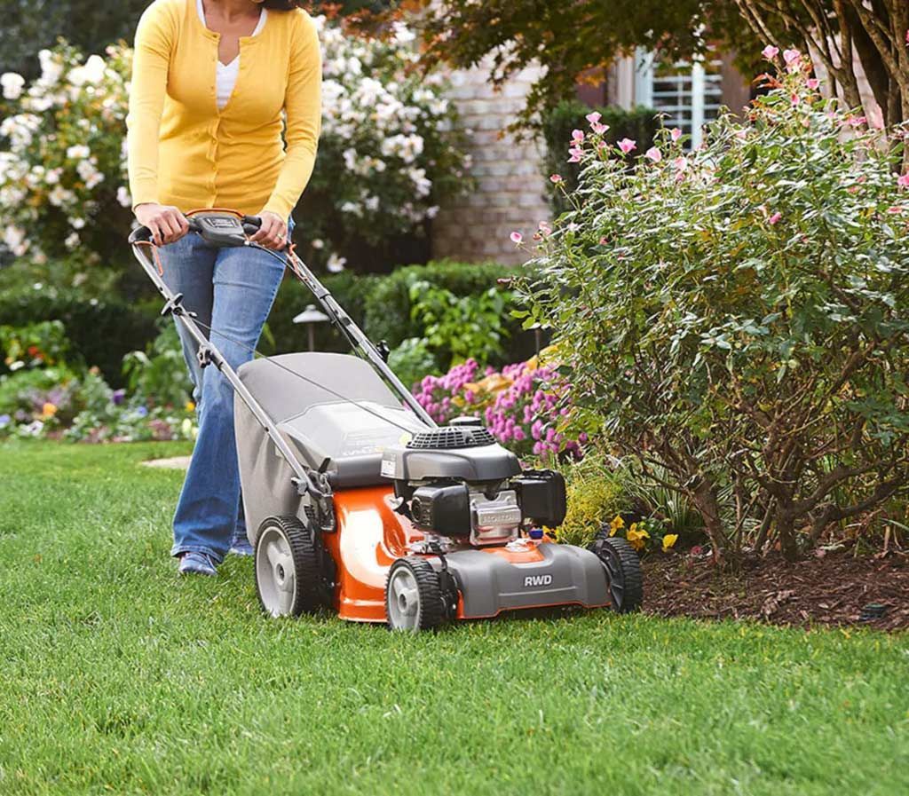 A Woman Is Pushing a Lawn Mower on A Lush Green Lawn — CBS Chainsaw & Brushcutter Specialists - HUSQVARNA in Harrington, NSW