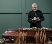 A man standing, reading from a book, with a casket adorned with flowers in the foreground, offering