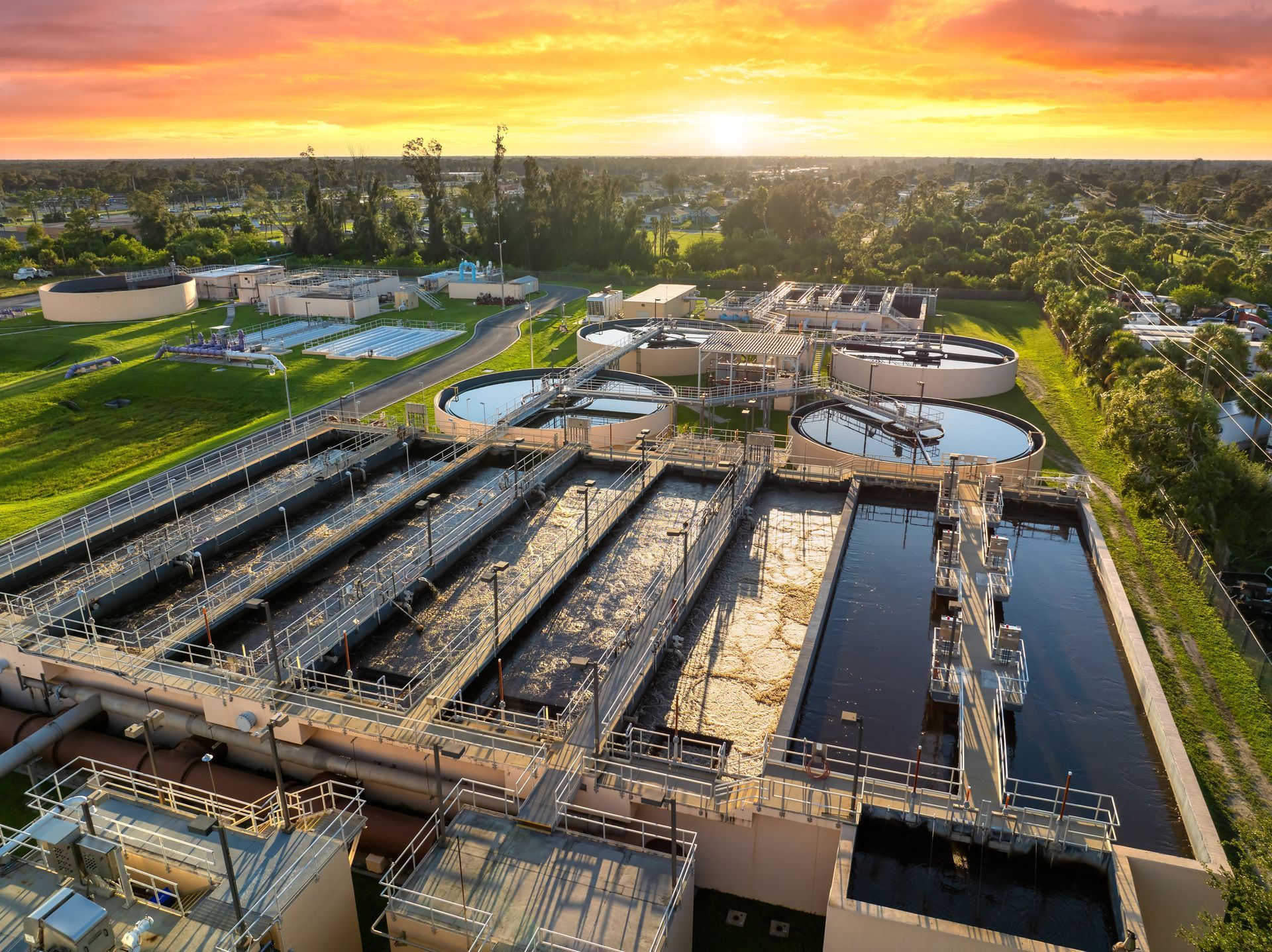 Aerial view of a wastewater treatment plant at sunset, with large tanks, pipes, and surrounding greenery.