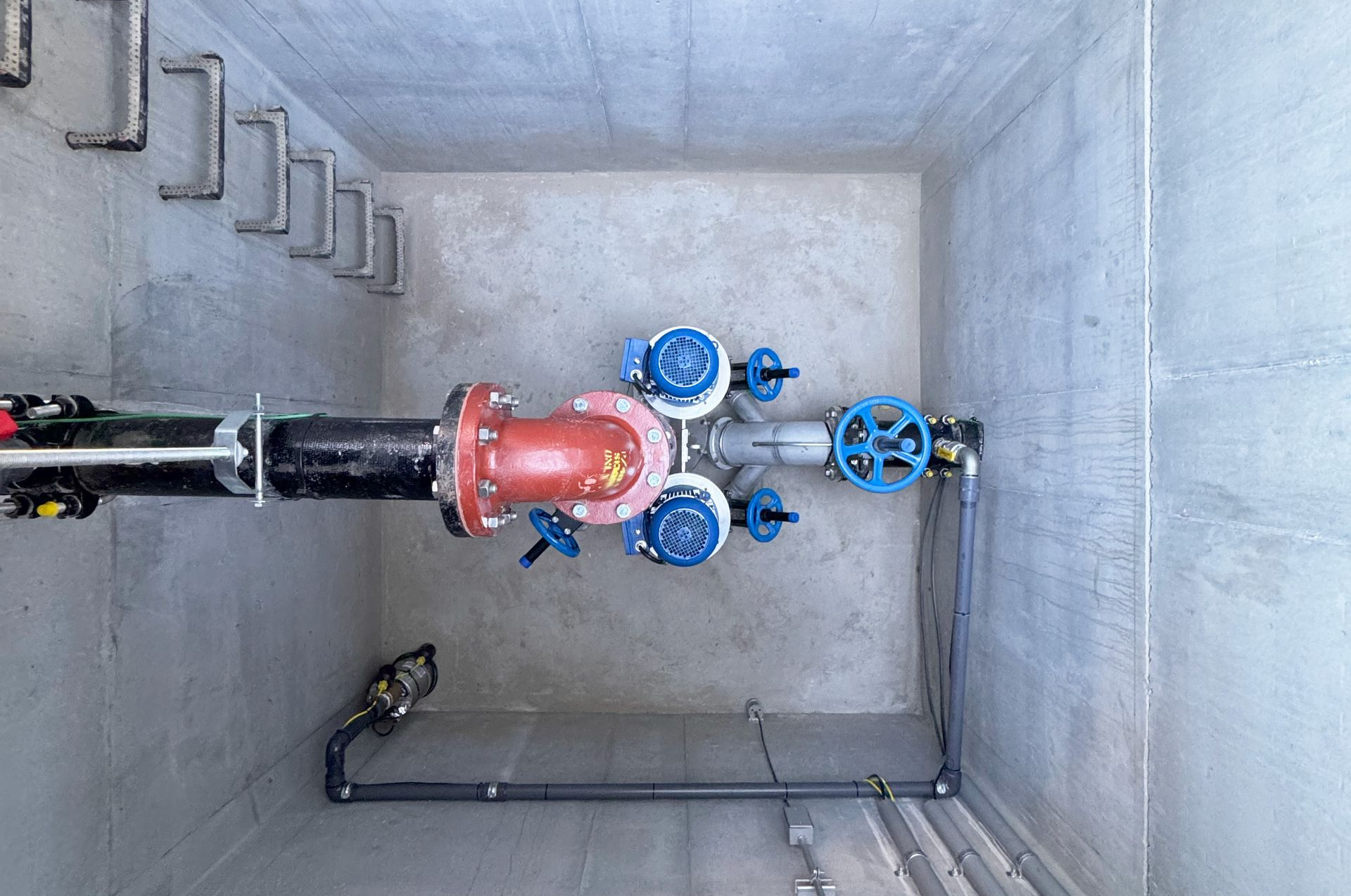 Industrial pipes and blue valves inside a concrete utility pit, with a large red pipe joint.