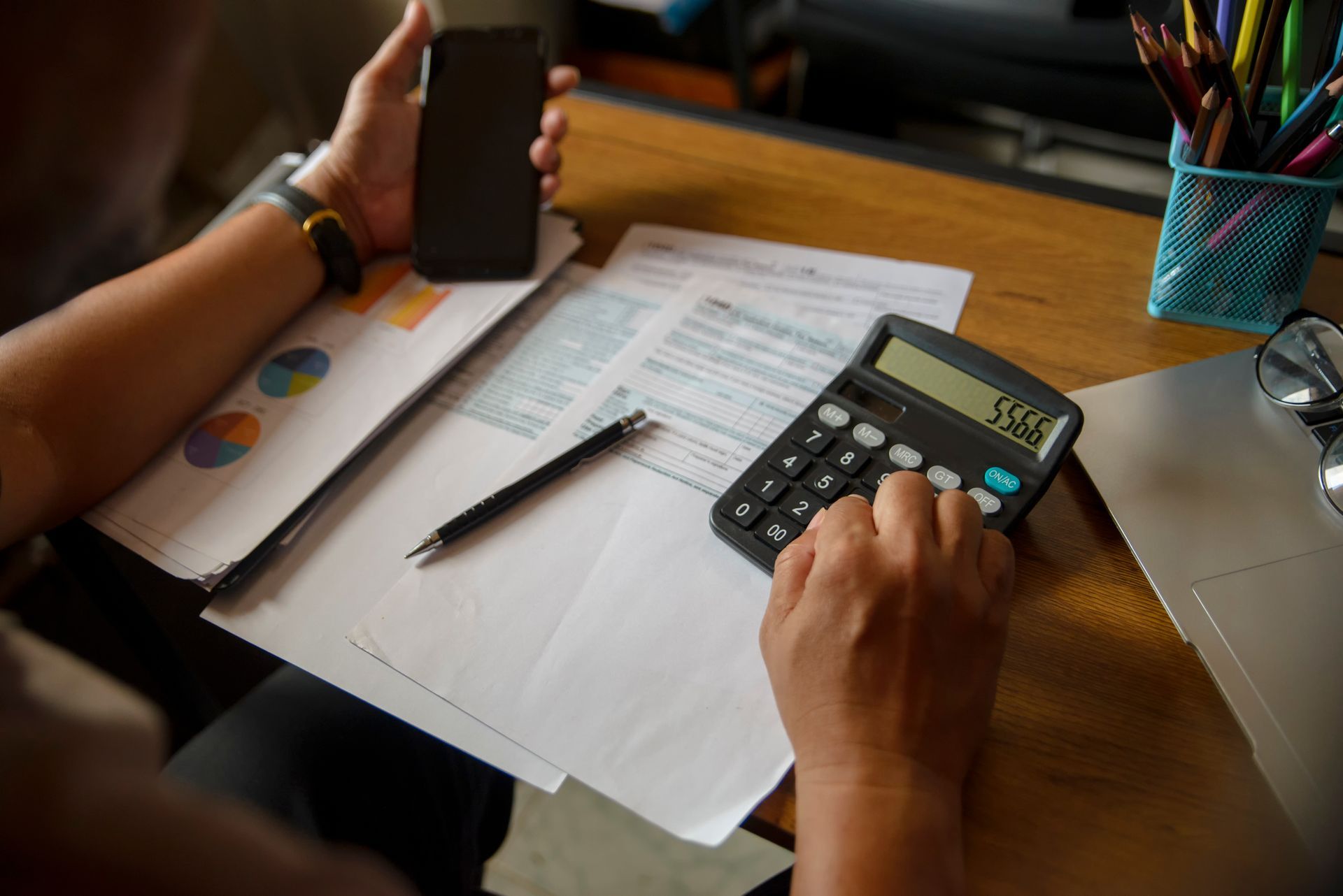 Person using a calculator, holding a phone, with documents and pen on a desk.