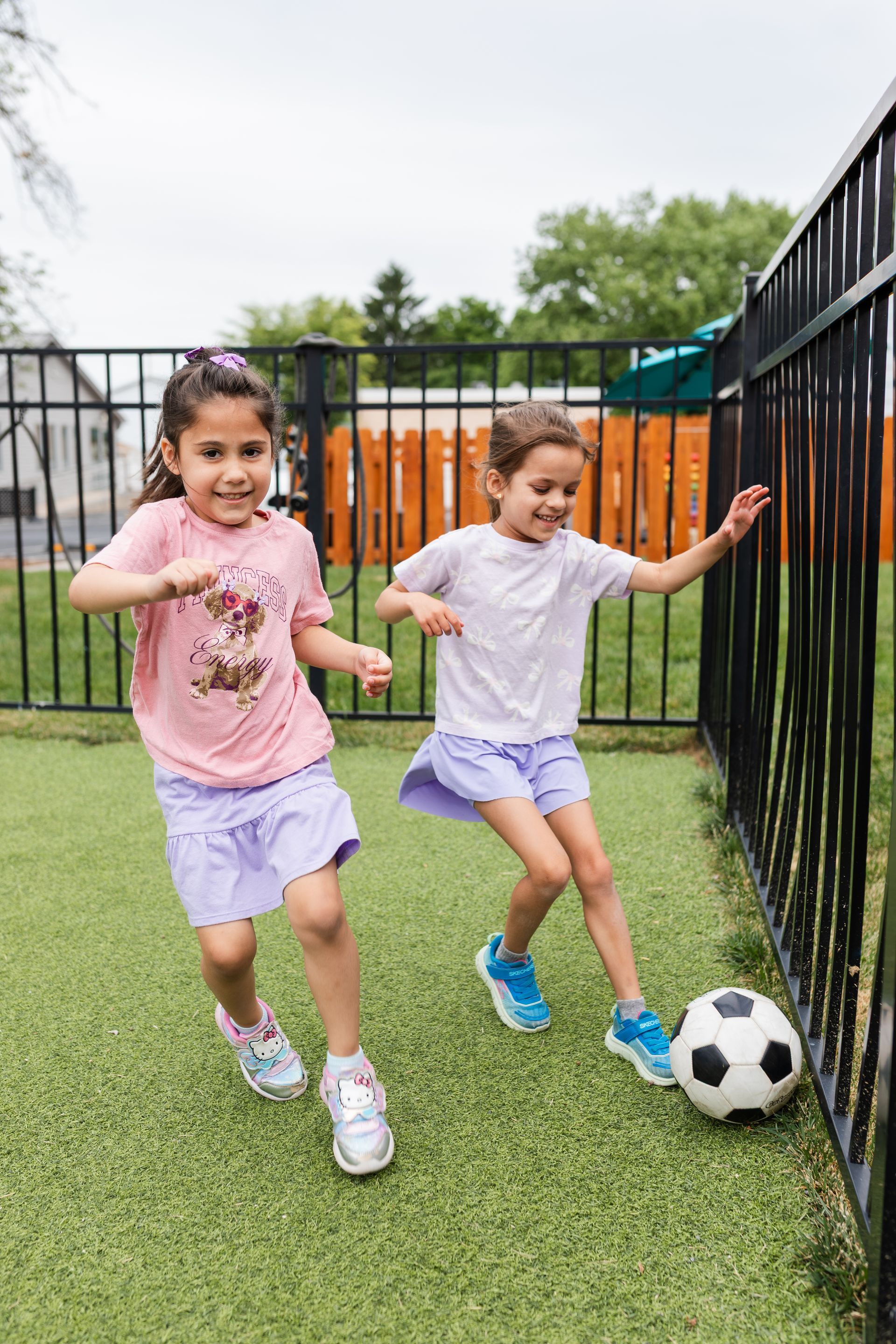 Two children playing soccer on grass beside a black fence, one smiling while the ball rests near the fence.
