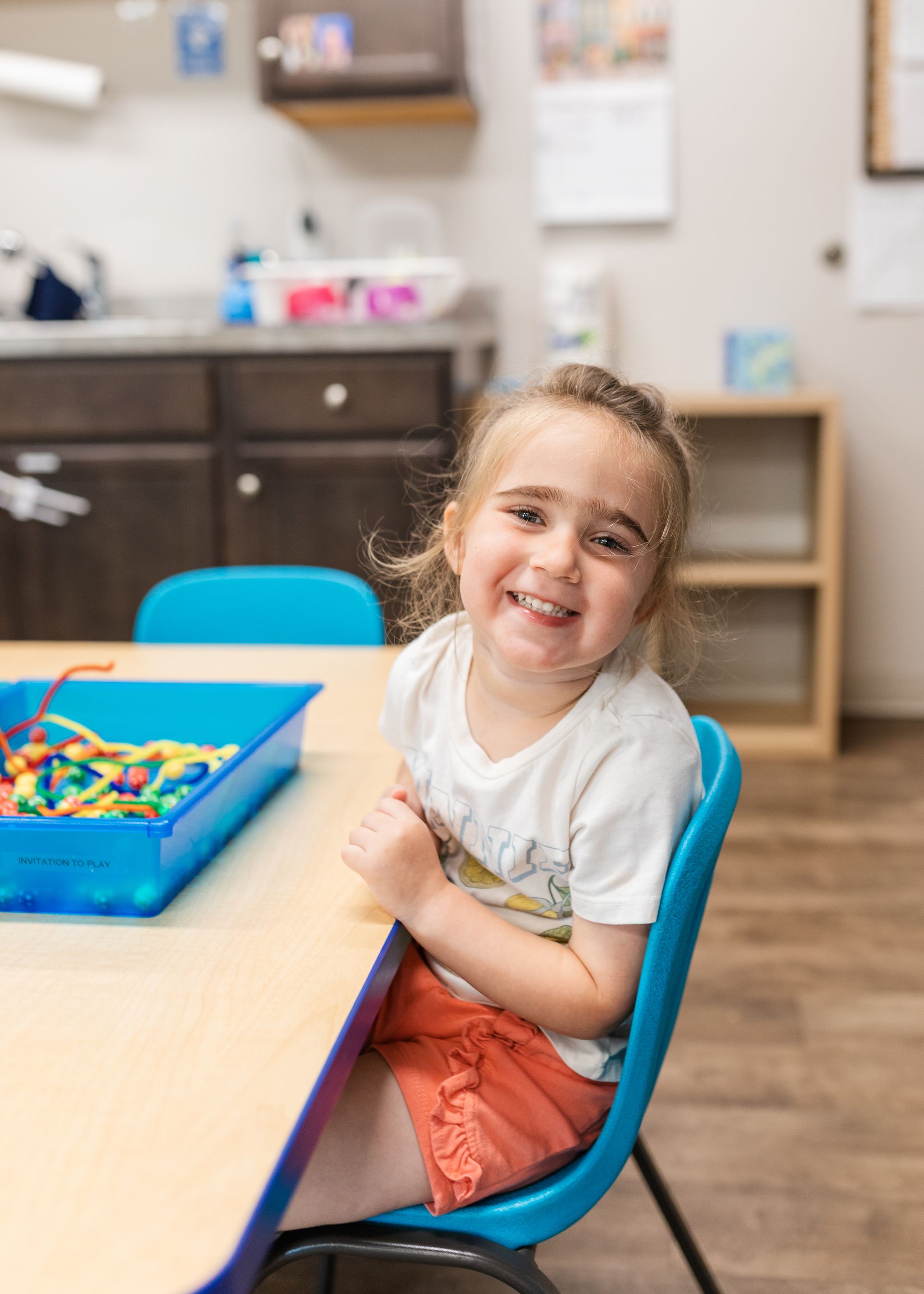 Smiling child seated at a classroom table with a blue chair and colorful activity tray