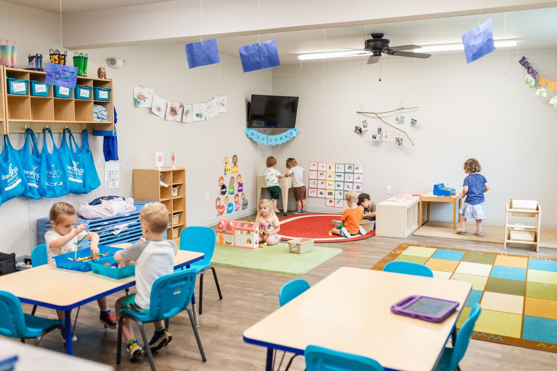Empty classroom with blue walls, desks, chairs, cubbies, and posters under bright fluorescent lights.