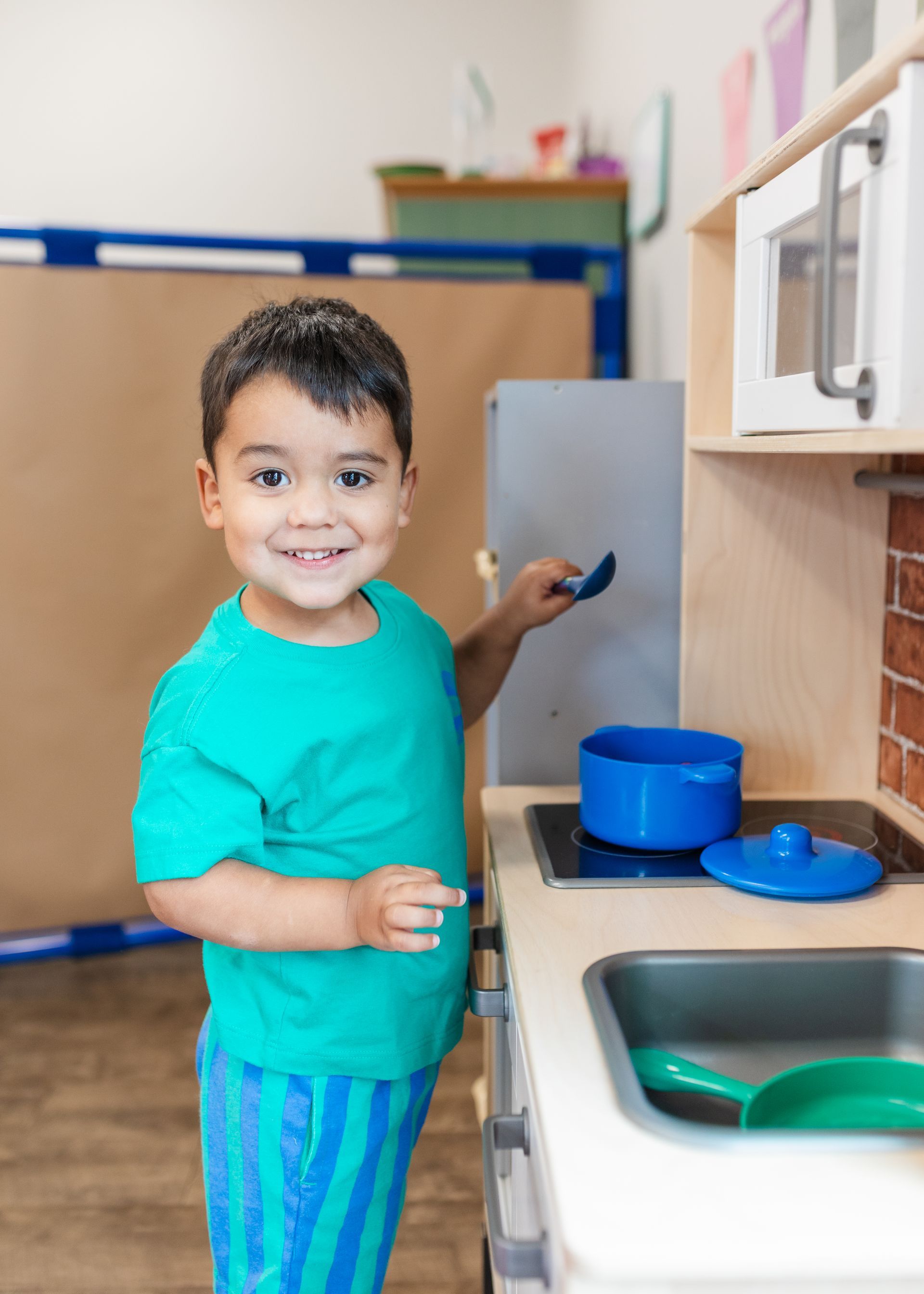 Child smiling in a play kitchen, holding a toy utensil beside a blue pot and sink.