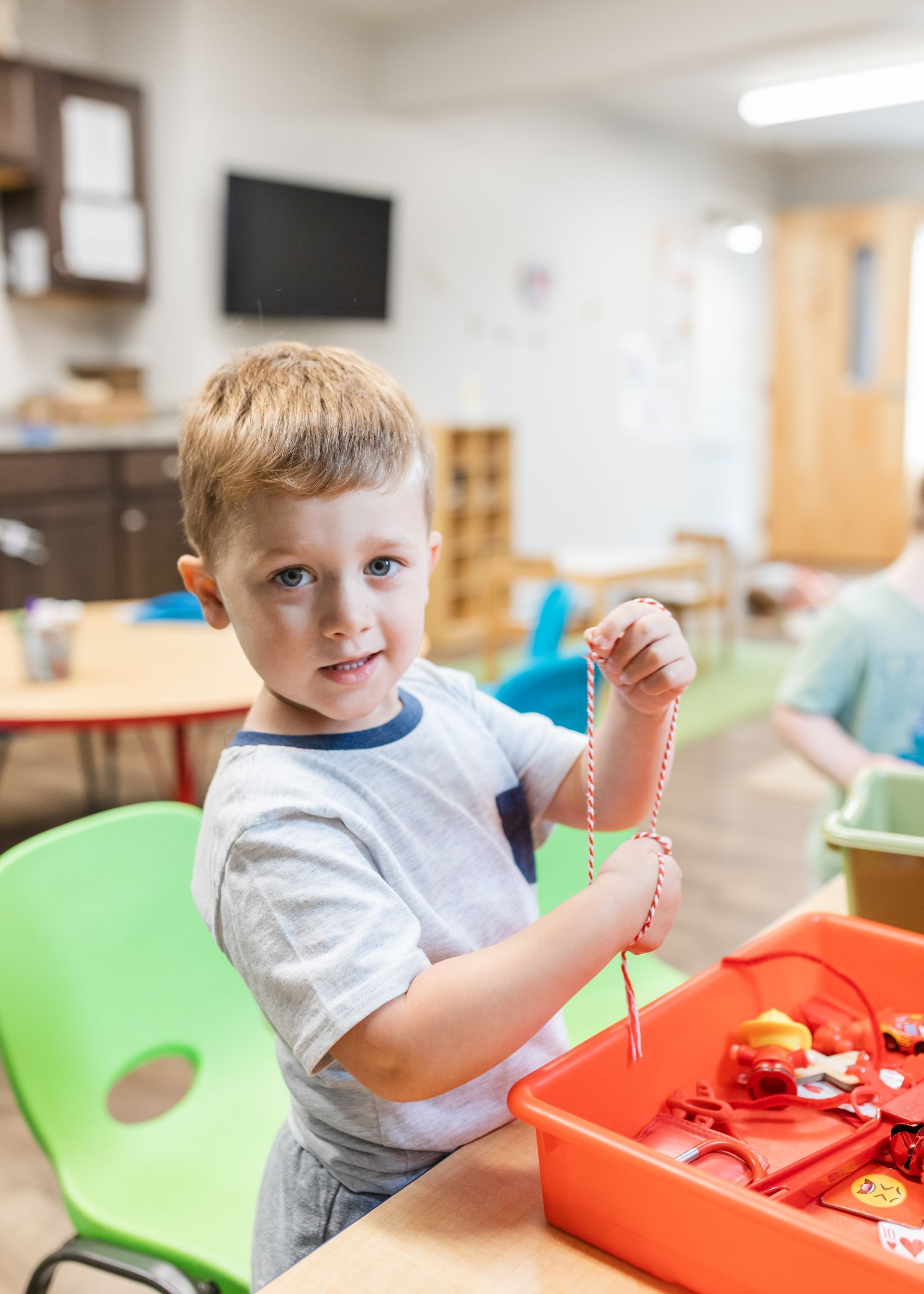 Child holding a beaded necklace next to an orange bin in a classroom, with green chairs and tables nearby