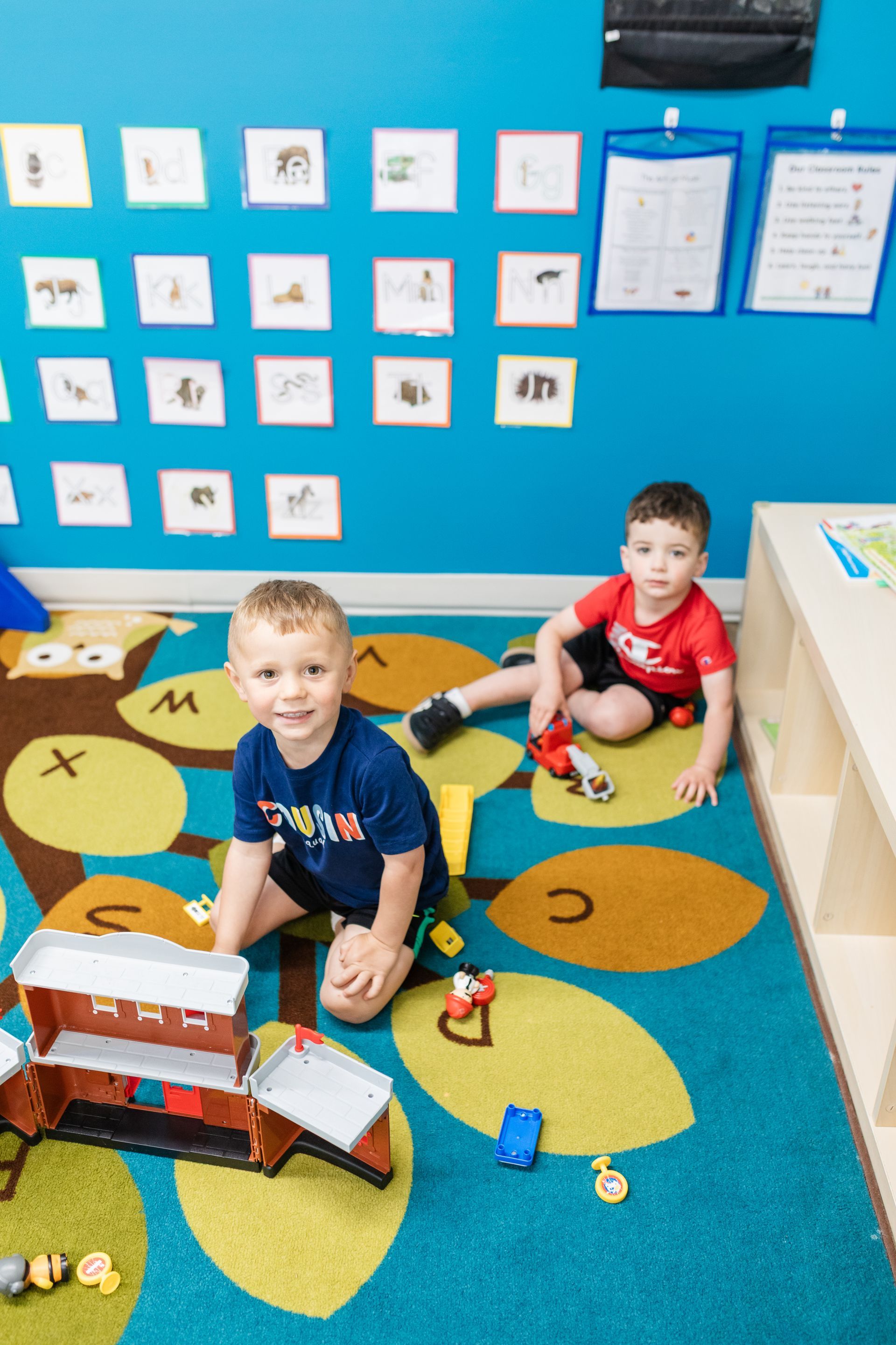 Two children playing on a colorful classroom rug with toys and wall displays behind them